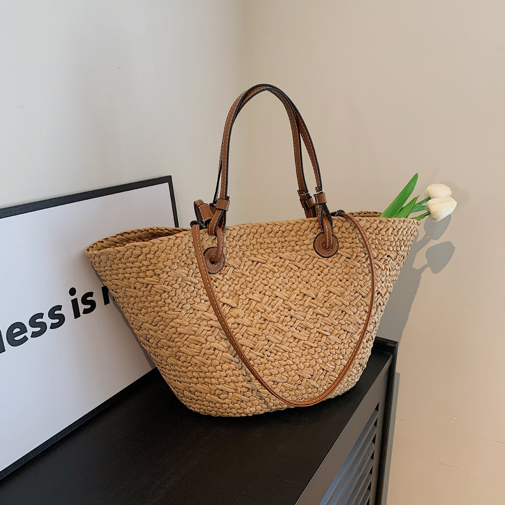 The Greemnest Artisan Paper Rope Woven Bag with brown leather handles rests on a black surface. White tulips peek out, and a nearby partial sign reading "ness is n" leans against the wall.