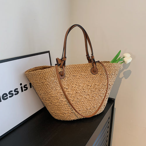 The Greemnest Artisan Paper Rope Woven Bag with brown leather handles rests on a black surface. White tulips peek out, and a nearby partial sign reading "ness is n" leans against the wall.