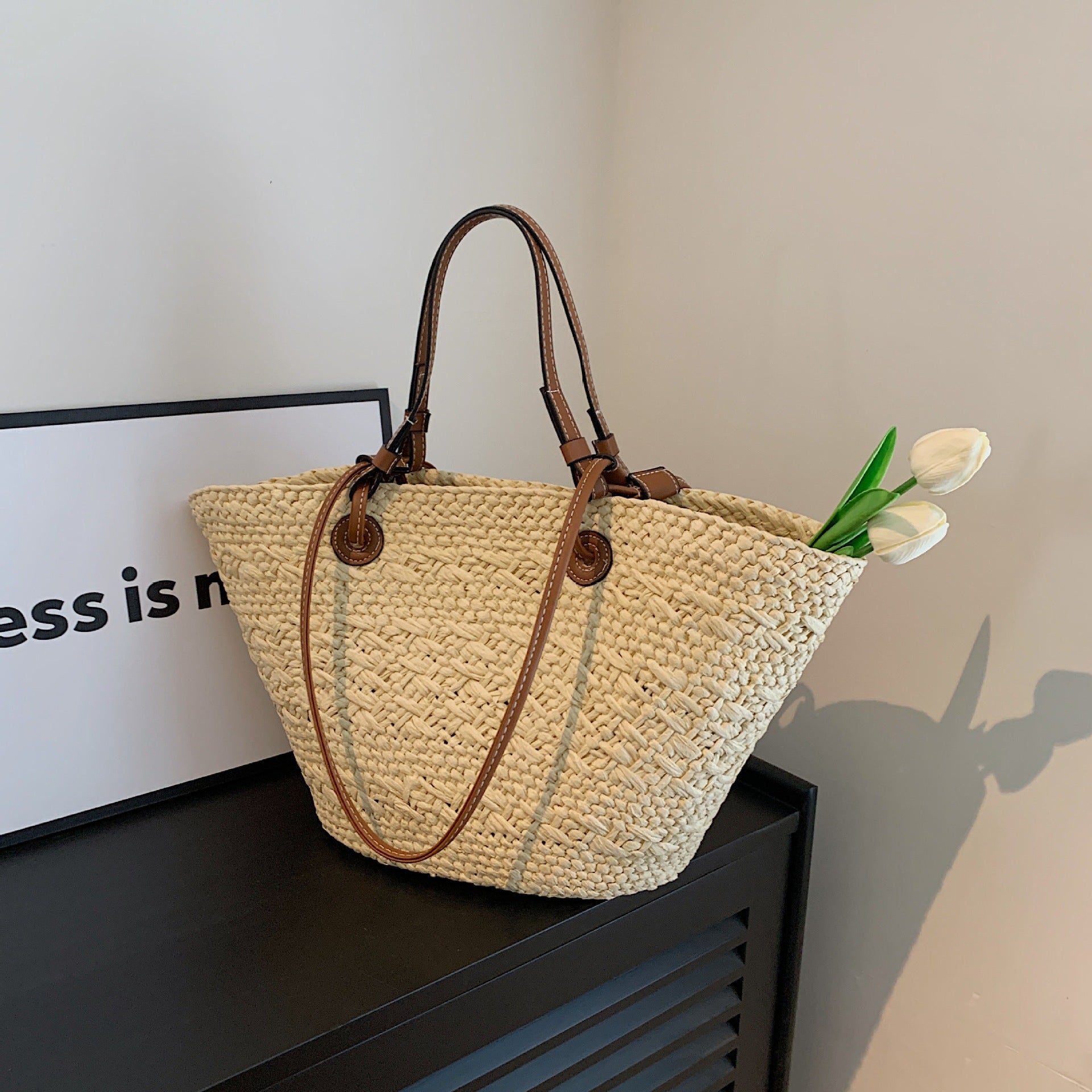 The Greemnest Artisan Paper Rope Woven Bag with drawstring closure and brown leather handles rests on a black surface. Two white tulips peek out from the eco-friendly bag, with part of a framed sign visible in the background.