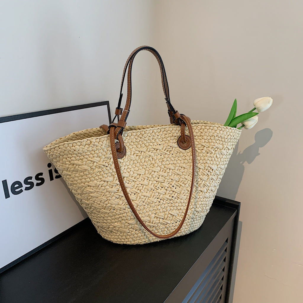 The Greemnest Artisan Paper Rope Woven Bag with drawstring closure and brown leather handles sits on a black surface, holding white tulips beside a framed sign partially displaying the words "less is.